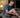 An older man sitting on a bed at night clutching his leg in pain with a bottle of medication nearby.
