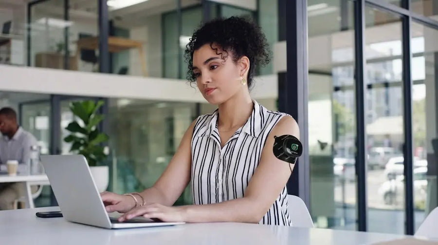 A woman is using a laptop at a modern office desk, wearing a black TENS unit on her arm.