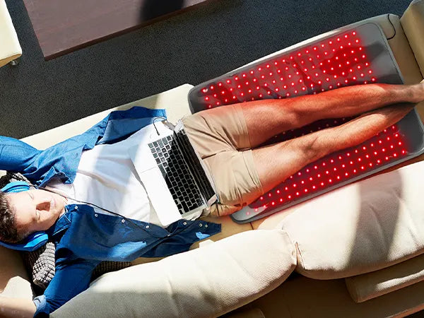 Man lying on a couch with a laptop, legs resting on a red LED light therapy mat.