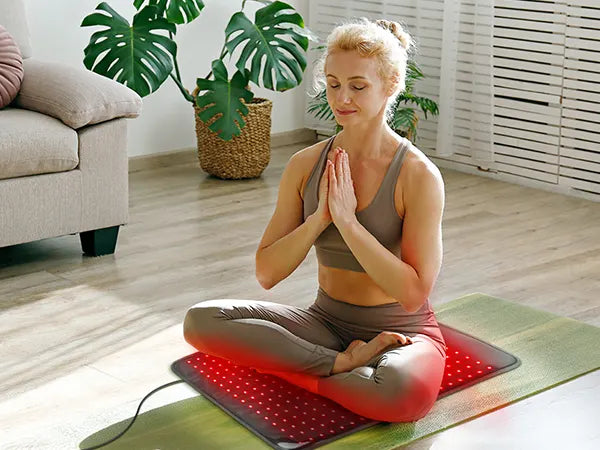 Woman meditating on a red infrared heating mat with a cord, sitting cross-legged in a bright room.