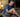 An older man sitting on a bed at night clutching his leg in pain with a bottle of medication nearby.