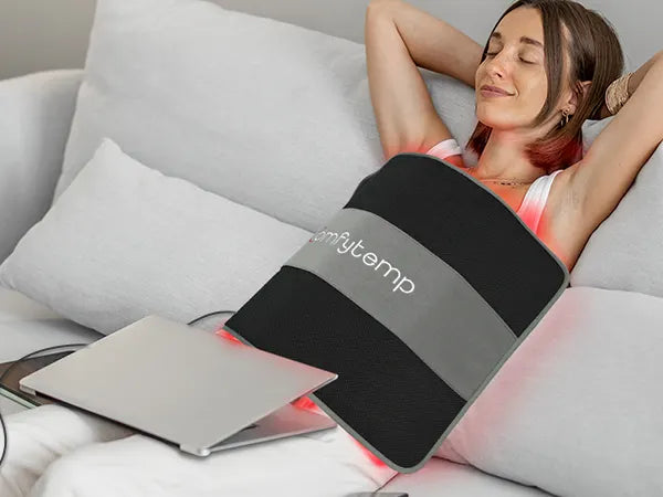 Woman relaxing on a sofa with a black and grey Comfytemp electric heating pad on her lap, emitting red light.