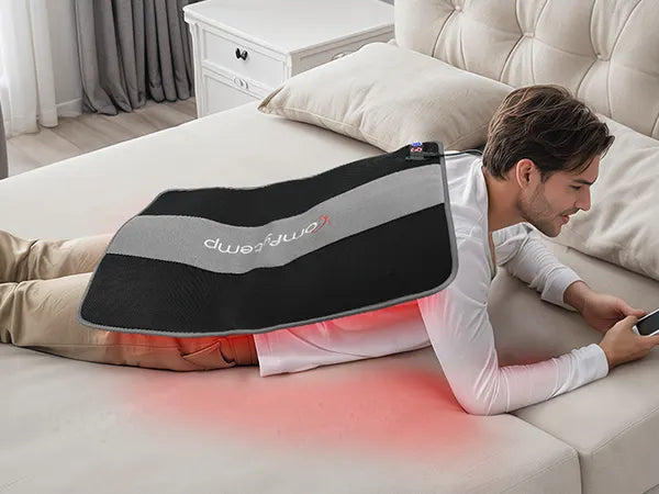 Man lying on bed using a black and grey Comfier heating pad with red light on his back.
