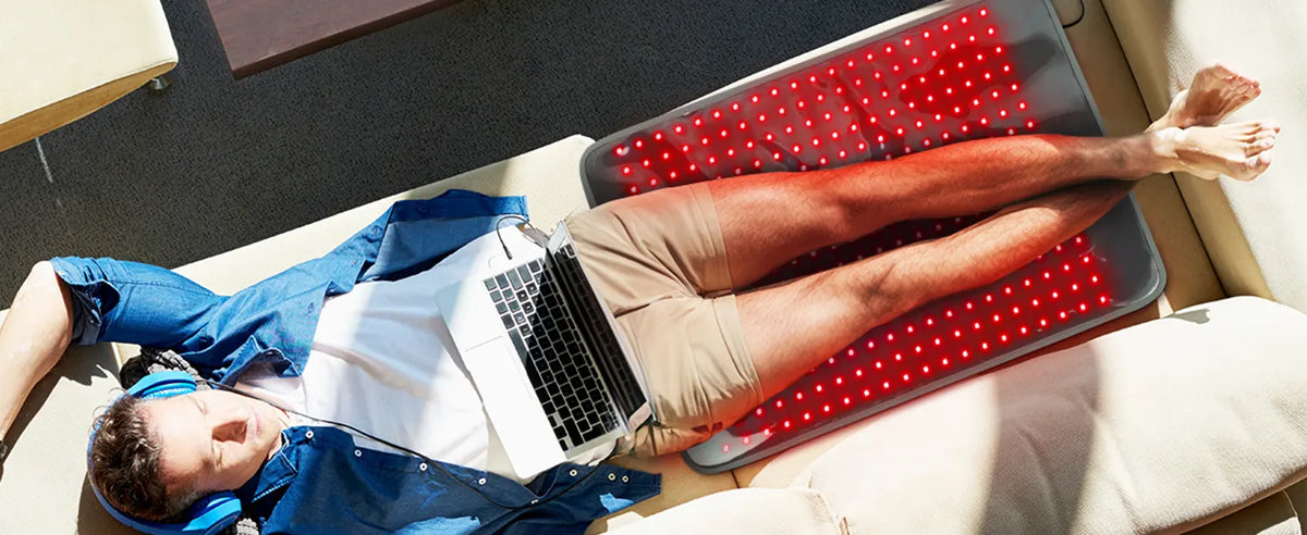 Man relaxing on a couch with a red light therapy mat glowing beneath his legs, wearing headphones and using a laptop.