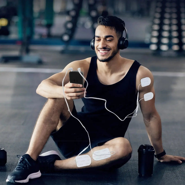 Smiling man in gym using TENS unit electrode pads on his arm and leg while looking at smartphone.