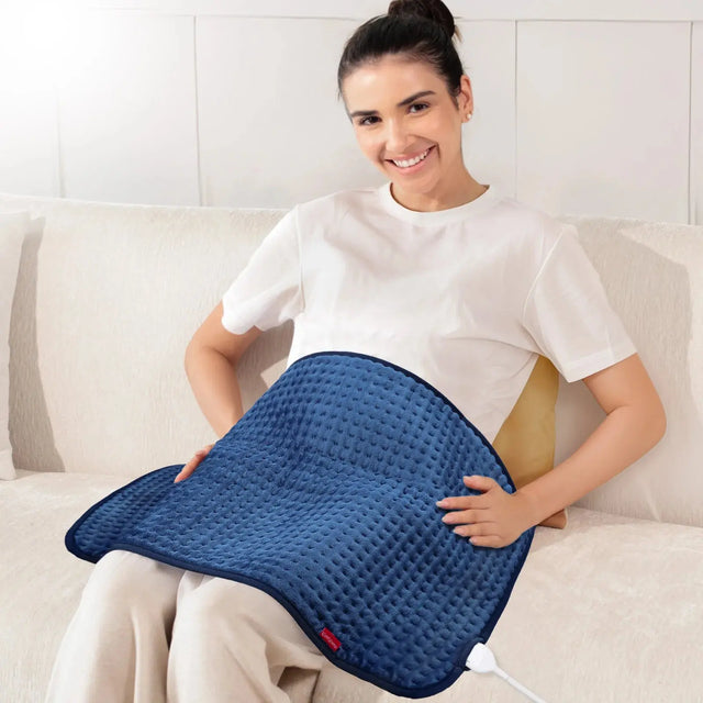 Smiling woman on a sofa using a blue Comfytemp electric heating pad on her lap.