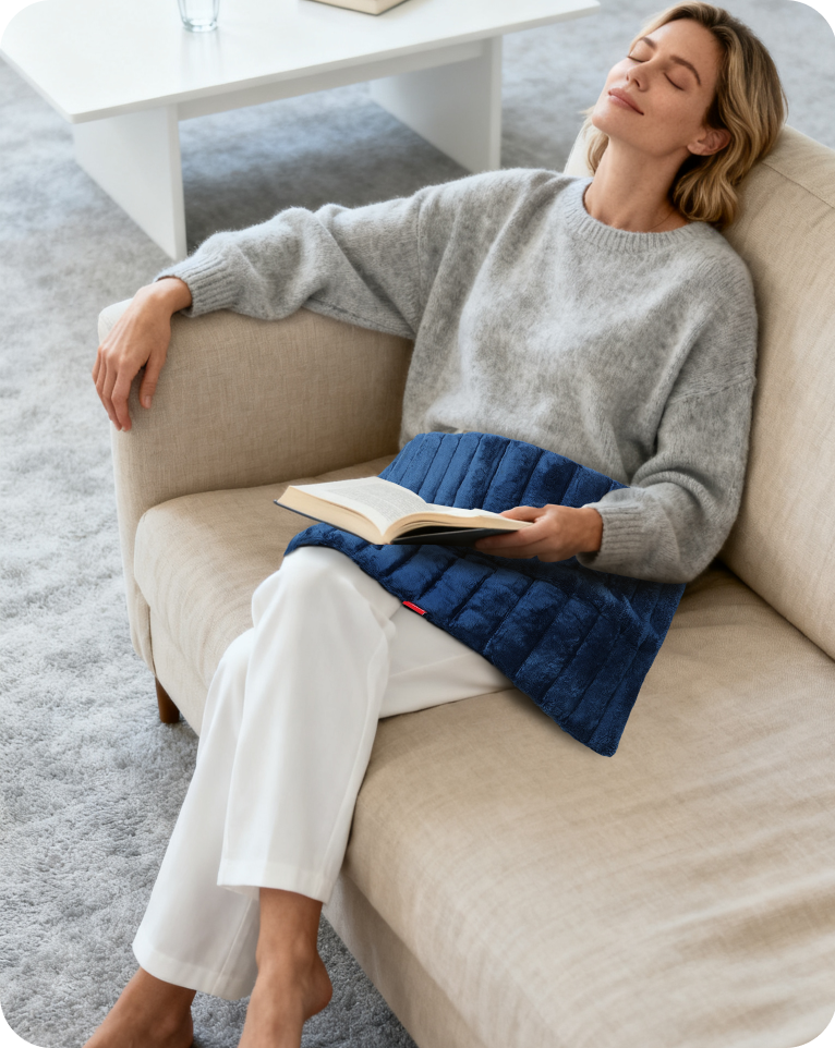 A woman relaxes on a sofa with a blue weighted heating pad on her lap while holding a book.