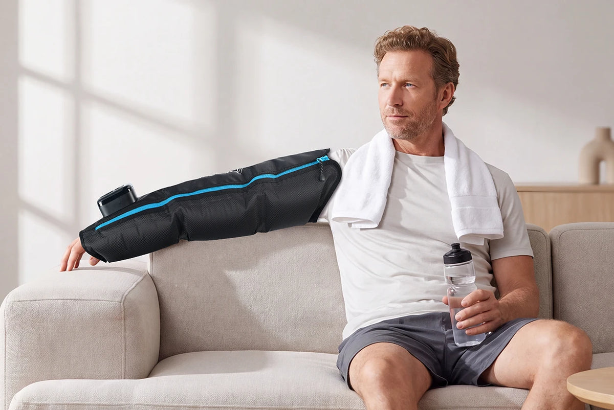 Middle-aged man on a beige sofa wearing a black arm compression sleeve with blue zipper, holding a water bottle.