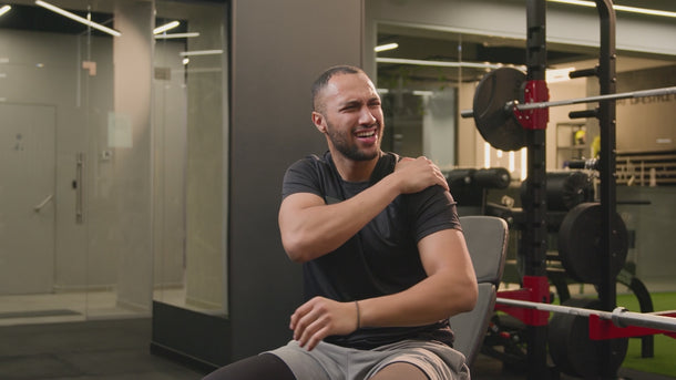 Man in a gym holding his shoulder in pain while sitting on a bench.