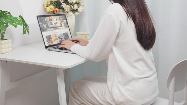 Woman in white long-sleeve top using a laptop on a white desk with plants and flowers nearby.